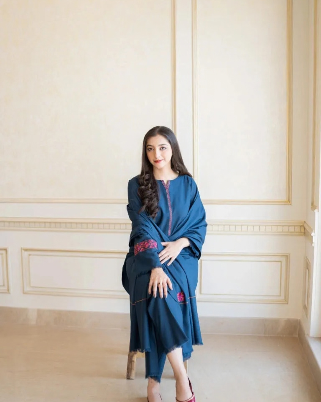 A woman wearing a dark teal blue traditional outfit with pink embroidered details sits on a wooden chair against a cream-colored paneled wall.